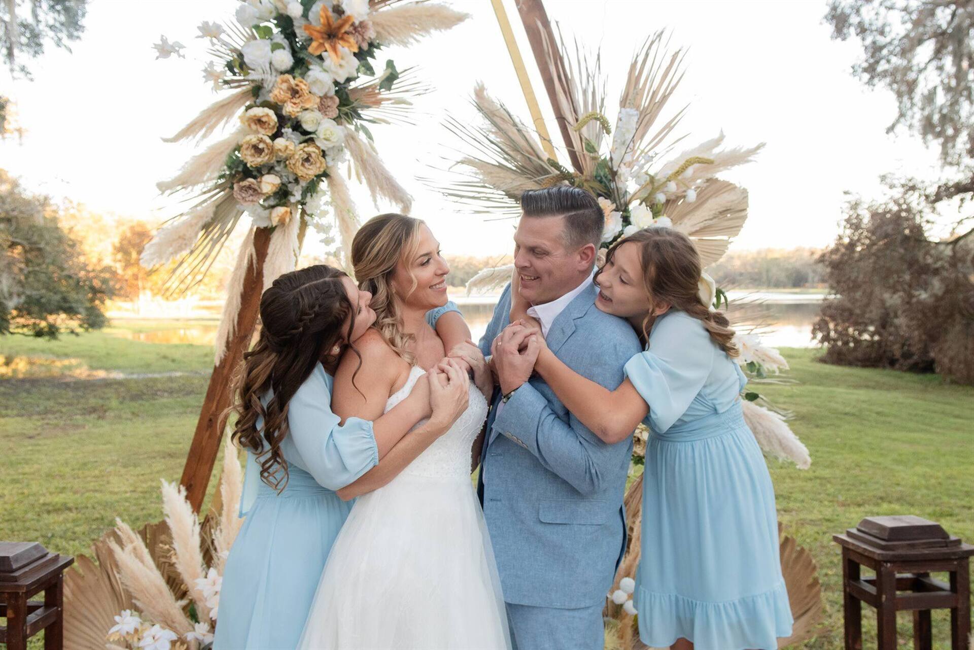 A bride and groom hugging two girls in light blue dresses in front of a floral arch by the lake at Lakeview Estate