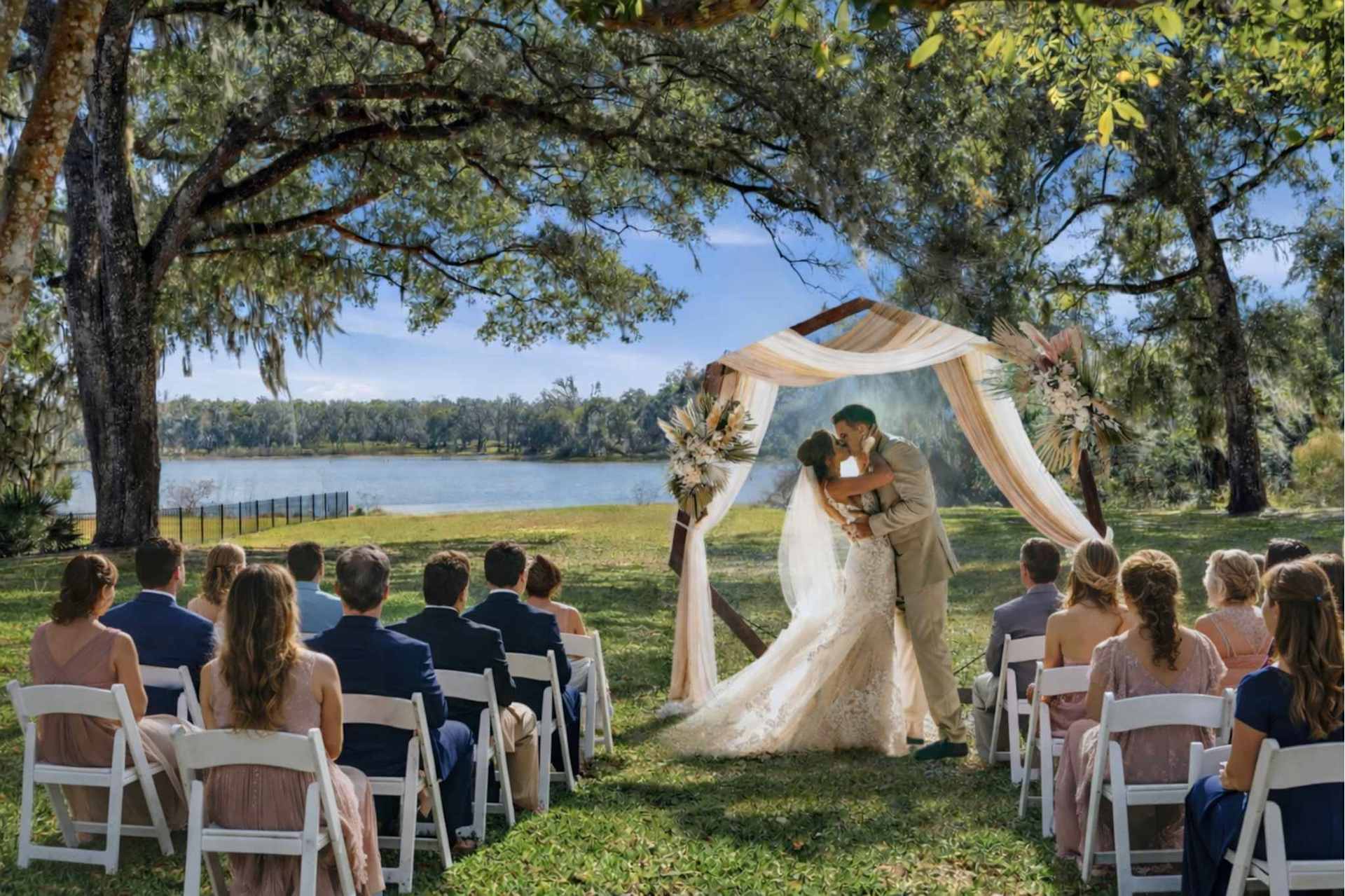 A ceremony setup idea on the estate lawn — guests in white chairs under the live oaks watch a couple kiss beneath a draped arch, with the lake behind them